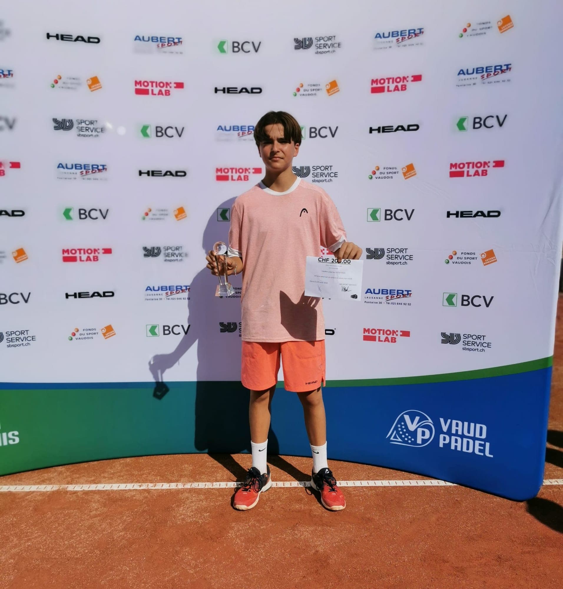 Lenart Kadriu avec sa coupe devant le mur des partenaires lors d’une remise de prix vaudoise